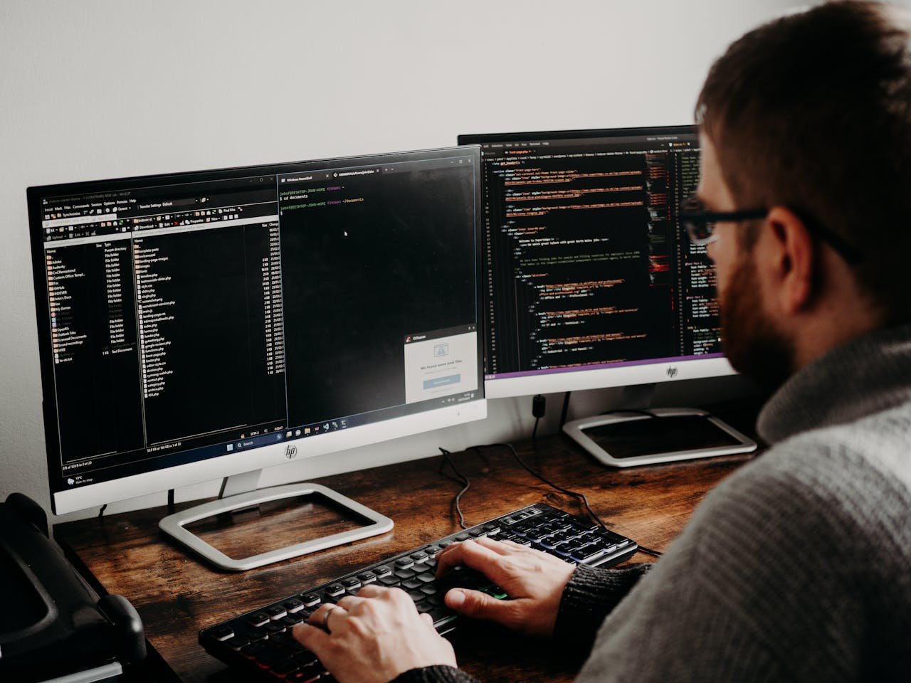 Software developer typing code on dual monitors at a wooden desk.