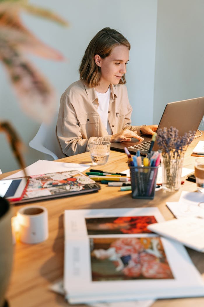 A young female designer works on a laptop in a creative workspace surrounded by art and design materials.